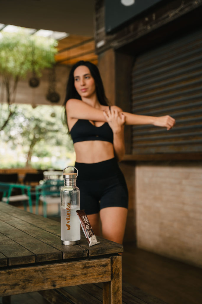 Imagem de mulher se alongando, vestindo roupas de prática de exercício físico e tendo hábitos saudáveis. À sua frente, uma garrafa de água e uma barrinha de proteína da vitanuts.